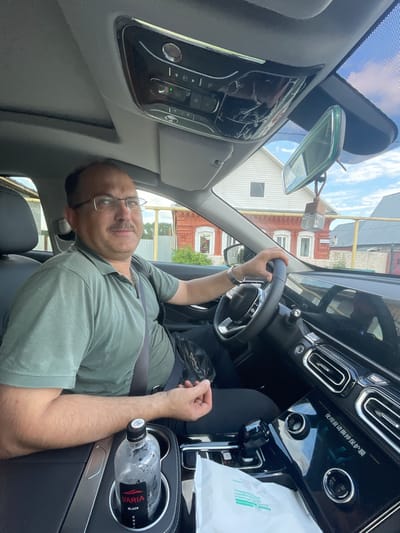 A middle-aged man with a mustache and glasses is seated in the driver's seat of a car, looking towards the camera. He is wearing a green polo shirt and a seatbelt. His right hand is on the steering wheel. The car's interior is visible, including the dashboard, rearview mirror, and a cup holder with a bottle of water. Outside the car, a rural scene with houses and a blue sky can be seen. The location is identified as Maminskoye, Russia.