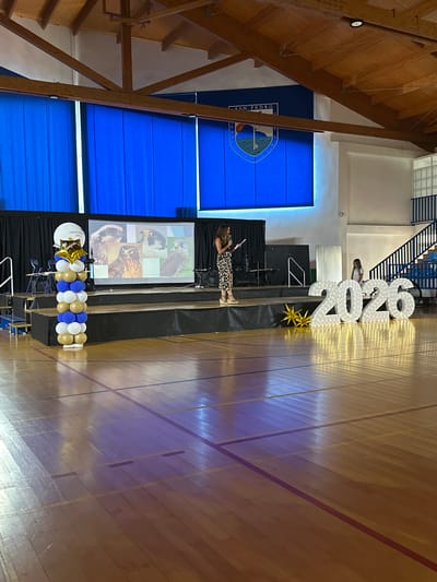 This media file captures an indoor event, likely a graduation ceremony, set in a gymnasium. The scene is dominated by a wooden floor with painted court lines and large, white, star-covered numbers spelling out "2026" as a prominent decoration. A woman in a patterned dress stands on a raised platform, holding a piece of paper and appearing to address an audience. Behind her, a screen displays images of birds, possibly falcons, in a grid format. To the left, a large blue banner with a crest and text, "SAN PEDRO," is visible, suggesting the location is associated with San Pedro De Alcántara, Spain. Another banner or backdrop, also blue, is present to the left of the screen. A cluster of gold and blue balloons, some shaped like stars, are arranged on the platform near the screen. The lighting suggests it is daytime or an indoor event with artificial lighting. The overall atmosphere seems celebratory, with the large numbers indicating a future graduation year. The presence of a stage, screen, and decorations points to a formal or semi-formal event.