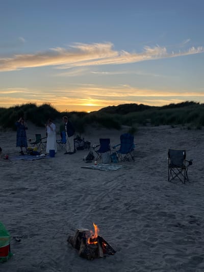 The image depicts a beach scene at dusk in Morfa Bychan, United Kingdom. A group of people are gathered on the sand, with a bonfire burning in the foreground. The sky is a gradient of blue and orange, with wispy clouds. Several folding chairs are set up, suggesting a picnic or gathering. In the mid-ground, three individuals stand near blankets and a cooler, while another person is seated in a chair on the right side of the frame. The background features sand dunes covered in grass.