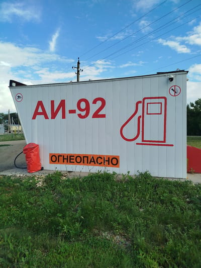 The image shows a white, modular building with red lettering that reads "АИ-92," indicating it is a gas station selling AI-92 gasoline. To the left of the lettering is a red, bag-like object with a hose attached, likely a fuel dispenser cover. On the wall, there is a red outline of a gas pump. Above the gas pump graphic is a circular sign with a diagonal red line through it and a stylized cigarette, signifying "no smoking." To the left of the "АИ-92" lettering, there is another circular sign with a camera icon inside a red circle, indicating surveillance. Below the "АИ-92" lettering is an orange rectangular sign with black lettering that reads "ОГНЕОПАСНО," meaning "flammable" or "fire hazard." The building is situated outdoors, with green grass and some gravel in the foreground. In the background, there are trees, a telephone pole with wires, and a cloudy blue sky. The scene appears to be in a rural or semi-rural setting, possibly on the outskirts of the city of Зиянчурино, Russia, given the provided context.