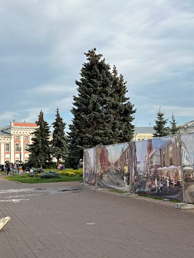 A street scene in Putyatino (Semivragovskaya S/A), Russia, is depicted under a cloudy sky. In the foreground, a paved area with some puddles leads to a large, colorful banner showcasing an artistic rendering of a European-style street with buildings, trees, and people. The banner is supported by metal frames and concrete blocks. Behind the banner, several tall evergreen trees stand in a grassy area. To the left of the trees is a building with red and white facade, featuring multiple windows and architectural details. A group of people are gathered near this building, some standing and conversing, while others are seated on the grass. A street lamp is visible to the left of the building. The overall impression is of a public space, possibly undergoing development or renovation, with artistic representations of the future appearance of the area.