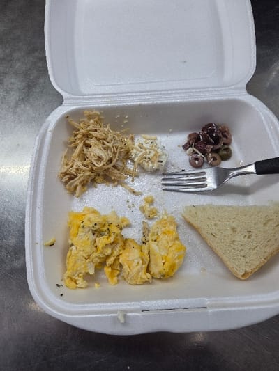An open white expanded polystyrene foam takeout container rests on a grey, reflective metallic surface. The container holds a multi-item meal. In the upper-left section, a portion of shredded light-colored meat is present. Adjacent to it, a small amount of white, granular dairy product with dark seasoning is observed. The upper-right section contains a cluster of sliced olives, comprising both dark and green varieties. A silver-colored metal fork with a black handle is positioned across the olive cluster. In the lower-left section, scrambled eggs, appearing partially disturbed, are present and seasoned with dark specks. The lower-right section contains a single triangular slice of white bread. The scene is located in Gevgelija, North Macedonia.