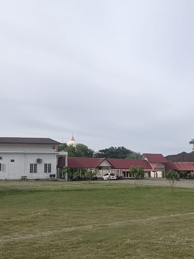 The image depicts a serene scene in Lampeudeu Baroh, Indonesia, under an overcast sky. In the foreground, a vast expanse of green grass stretches across a gently sloping field, its texture varied with patches of dry earth and small stones. Beyond the field, a low-lying building with white walls and a dark-tiled roof occupies the left side of the frame. It features multiple windows with white frames and a visible air conditioning unit. Adjacent to this building and extending to the right are several structures with distinctive red, sloped roofs. These buildings appear to be part of a complex, possibly a school or a communal area. Two small, leafy trees stand prominently in front of these red-roofed buildings, adding a touch of greenery to the architectural landscape. Parked vehicles, including motorcycles and a white car, are visible near the entrance of one of the buildings. In the distance, a cluster of trees forms a dense green backdrop, partially obscuring a distinctive golden dome, a possible religious or cultural landmark. The overall impression is one of tranquility and everyday life in a semi-urban or rural setting in Indonesia.