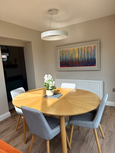 The image captures a modern dining area within a home in Harrogate, United Kingdom, bathed in bright, natural daytime light. The central feature is a round, light wooden dining table with a sunburst pattern radiating from its center. Four modern dining chairs with light grey fabric upholstery and wooden legs are arranged around the table; three are fully visible, and the cushion of a fourth is partially seen underneath.

On the table, a small potted plant with white flowers adds a touch of greenery, positioned next to a grey book or notebook. The flooring throughout the space is a light brown wood-effect laminate or engineered wood.

Against a light, neutral-toned wall, a striking rectangular artwork is prominently displayed, framed in a light grey or white. The painting features an abstract depiction of a forest, characterized by vibrant vertical streaks of yellow, orange, and blue, overlaid with black silhouettes of tree trunks and branches. Below the painting, a white, slatted radiator is mounted on the wall.

A contemporary, circular white LED pendant light fixture hangs from the ceiling, connected to a circular base on the wall by thin wires. To the right of the table, a partial view of a fifth chair or cushion with orange fabric is visible. Through a doorway in the background, a glimpse into another room reveals a darker interior with what appears to be a dark sofa and a circular mirror on a wall. There is no visible text.