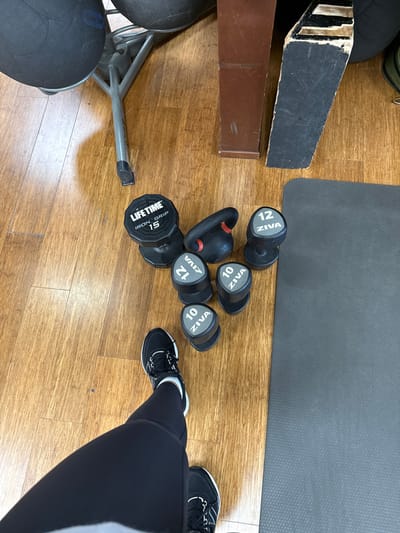 An overhead view captures a scene on a light brown wood-laminate floor in Plainsboro Township, United States. In the bottom left, the lower legs and feet of an individual are visible, clad in black athletic leggings and black athletic shoes with white soles and laces. A collection of exercise equipment is arranged on the floor: one black "LIFETIME IRON GRIP 15" dumbbell, one black kettlebell with a red accent, two black "ZIVA 12" dumbbells, and two black "ZIVA 10" dumbbells. A dark grey exercise mat occupies the right side of the frame. In the background, partially visible objects include a silver-colored metal stand, a brown wooden furniture element, and a dark, irregularly shaped object.