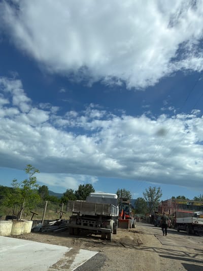 The image depicts a scene in Zlati Voyvoda, Bulgaria. A clear blue sky with scattered white clouds occupies a significant portion of the upper-left area. Several power lines are strung across the upper portion. On the right side, there's an industrial yard with various vehicles, including trucks and a tractor. An individual is standing beside some vehicles. A mix of trees and vegetation is also present. The ground is mostly dirt and gravel, with some concrete slabs.