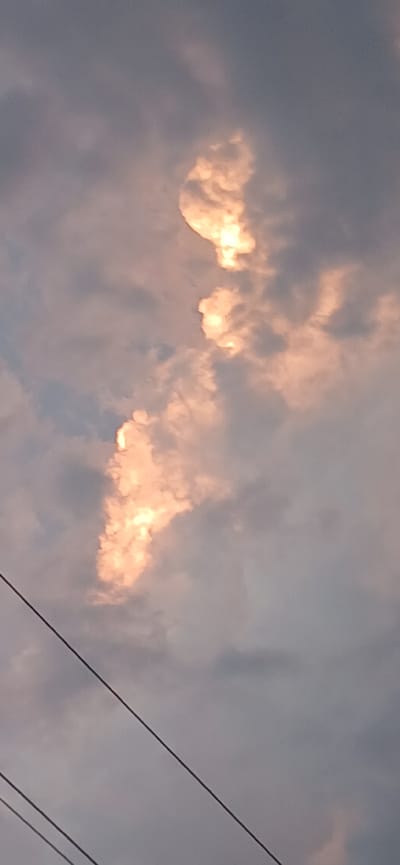 The image depicts a sky filled with clouds. Some of the clouds are illuminated by a warm, golden light, suggesting that the sun is either rising or setting. The light creates a dramatic effect, with some cloud formations appearing brightly lit while others remain in shadow, giving them a textured, almost sculpted appearance. The overall color palette is muted, with shades of grey, blue, and soft orange. In the foreground, two dark, thin wires stretch diagonally across the frame from the right side. These wires are likely power lines or telephone wires, indicating an urban or semi-urban setting. The presence of clouds suggests the weather is either overcast or transitioning. The scene is likely captured outdoors during twilight. No people or specific landmarks are visible, and there is no visible text in the image.