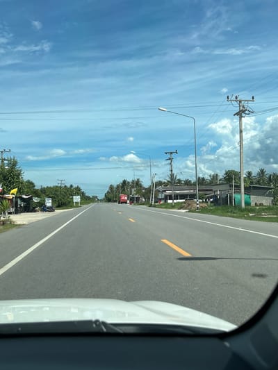 The media file depicts a roadside scene in Ban Thung Suea Non, Thailand. The perspective suggests the viewer is inside a vehicle, looking out at the passing scenery. The road, paved with asphalt, has a white line marking its edge and a broken yellow line indicating the center. The right side of the road is bordered by a grassy shoulder and some vegetation.

To the left of the road, a series of utility poles with electrical wires stretches into the distance, interspersed with streetlights. Beyond the poles, a few buildings are visible, appearing to be shops or residences, with roofs of varying materials. Palm trees are abundant, indicating a tropical environment. The sky is a bright blue, with scattered white clouds.

Further down the road, more buildings and trees line the landscape. Several white signs with text in Thai are visible along the side of the road. A red truck is seen on the road in the distance. The overall atmosphere is one of a bright, sunny day, typical of a rural or suburban setting in Thailand. The lighting suggests it is daytime, likely mid-morning or early afternoon. No people are prominently featured, but there is a hint of what might be a parked motorcycle on the side. The scene conveys a sense of travel and everyday life in a tropical location.