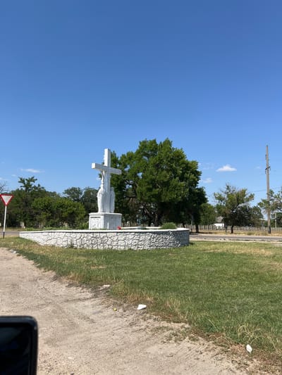 The image shows a white statue of Jesus Christ on a cross, with a tombstone behind it. The statue is located in a graveyard in Butor, Moldova. The sky is blue and clear, indicating a sunny day. The grass is green, and there are trees in the background. A road is visible in the distance, with a power line running parallel to it. The overall scene suggests a peaceful and solemn atmosphere. The statue is detailed, with Jesus's body and the cross clearly defined. The tombstone is also elaborate, with carvings and inscriptions. The graveyard appears to be well-maintained, with neatly trimmed grass and flowers. The presence of trees adds to the natural beauty of the setting. The road and power line suggest that the graveyard is located near a populated area, but it still retains a sense of tranquility.