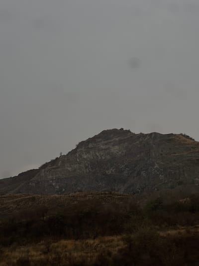 The image depicts a mountainside, presumably near San Bernardino Chalchihuapan, Mexico, under an overcast sky. The left portion of the frame is filled with a gray, featureless sky, while the right side showcases the steep, rugged terrain of the mountain. The mountain's surface is varied, exhibiting areas of dark, rocky cliffs alongside patches of brown vegetation. The overall environment appears harsh and potentially remote. There are no people, animals, or man-made structures visible in the image.