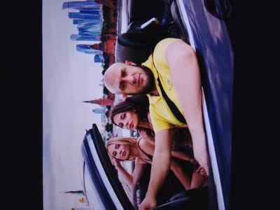 The image shows three people in a convertible car, with a city skyline in the background. The man in the driver's seat is bald with a beard and is wearing a yellow polo shirt. Two women are in the passenger seats. The woman in the middle has long dark hair and is looking at the camera. The woman in the front passenger seat has blonde hair and is looking down. The city skyline appears to be Moscow, Russia, given the onion-domed churches and modern skyscrapers. The sky is overcast, suggesting it might be daytime. The overall mood of the image is casual and relaxed. There is some text visible on the man's shirt, which appears to say "MONCLER".