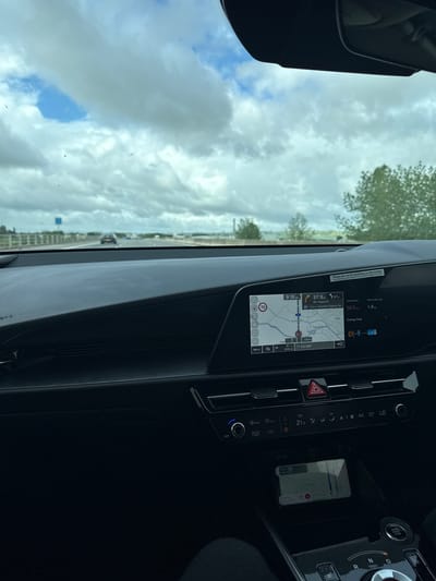 The media file captures the interior of a modern car, likely in the United Kingdom, looking out through the front windshield. The car appears to be traveling on a highway under a cloudy sky with some patches of blue.

The dashboard features a large, integrated digital display unit. The left screen shows a navigation map with a route highlighted. Visible text on this screen includes the current time "9:19 AM", current speed limit "70" mph, a warning label "Please do not fix anything to this screen. Any damage could result in a charge of £2,500", and navigation details: "37.9 mi" to MS Plymouth, "12.2 mi" to Taunton Deane Service area, and an estimated arrival time of "10:03 AM". Specific landmarks like "Hamp Bridge" and a postcode "EX2 8DP" are also shown on the map, consistent with a location near North Petherton, UK. The display also indicates a "Fuel economy" of "50.5 mpg" and an "Energy flow" graphic, suggesting a fuel-efficient or hybrid vehicle.

To the right of the infotainment screen are climate control settings, showing a temperature of "21.5" degrees Celsius, and buttons for "AUTO CLIMATE" and "PWR". A red triangular hazard light button is prominent. Further right, a rotary gear selector displays "P, R, N, D" positions, next to an "ENGINE START STOP" button. In a wireless charging tray below the climate controls, a smartphone is visible, also displaying a navigation map with "Stay on M5" shown, reinforcing the vehicle's activity of being in transit.

Outside the car, a road or highway is visible, with a distant vehicle and trees along the roadside. The overall scene depicts a morning journey by car.