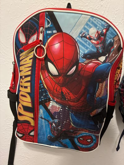A Spiderman backpack is hanging on a white wall. The backpack features Spiderman in various poses, with the words "SPIDER-MAN" printed vertically on the left side. There is a circular emblem on the bottom right of the backpack with Spiderman's mask and the text "WITH GREAT POWER COMES GREAT RESPONSIBILITY". The backpack has red trim and zippers.