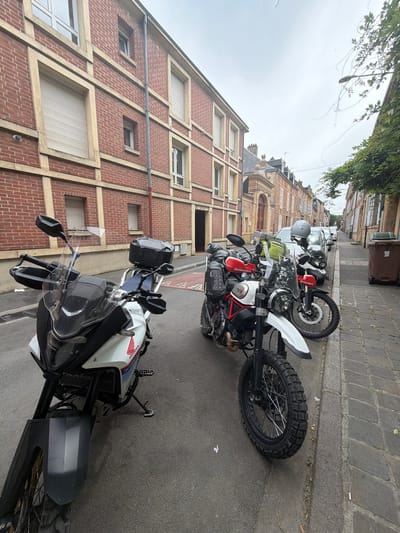 On a cloudy day in Charleville-Mézières, France, several motorcycles are parked along a cobblestone street, adjacent to a sidewalk. The front motorcycle is a white and black Honda with a windshield and black luggage mounted on the rear. Behind the Honda are additional motorcycles of varying colors, including a white and black model with red accents, a gray helmet resting on the seat, and another further back with a red fuel tank. The motorcycles are parked in front of brick buildings with stone accents and closed window blinds. A green trash bin is visible next to the sidewalk.