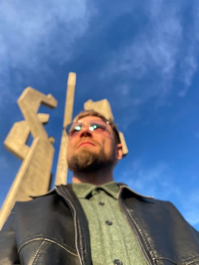 An adult male is depicted in a low-angle photograph, positioned in the foreground and looking upwards and slightly to the viewer's right. He has light brown hair, a short beard, and a mustache, and is wearing sunglasses with reflective lenses displaying blue and pink hues. His attire includes a collared, light green button-down shirt, partially visible beneath an open, dark, possibly leather jacket with a zippered front and white stitching details on the shoulders. The background consists of a clear blue sky with sparse, wispy white clouds. In the out-of-focus mid-ground, a large, Brutalist-style concrete monument, identified as the Monument to the Fallen Fighters, stands against the sky in Barutana, Montenegro. The scene is illuminated by direct sunlight, creating bright reflections on the man's sunglasses and highlighting portions of the monument.