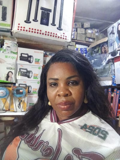 A medium close-up shot depicts a woman in an indoor commercial setting in Akodo, Nigeria. The woman, of African descent, occupies the center-left foreground, facing the camera with a neutral expression. She wears a white, mesh-textured short-sleeved top featuring maroon/brown graphics and a green "ASOS" logo on the upper right chest. Her long, dark hair is styled down, and she has visible makeup around her eyes and on her lips. Gold-colored, teardrop-shaped earrings are present on both ears.

The background is densely packed with merchandise, indicative of a retail establishment. Immediately behind the woman, stacked white boxes display images of black, rectangular "NEW POWER" brand flashlights/torches, some marked "LED" and "rechargeable." Below these, two blue, circular solar-powered lights with stands are visible in clear packaging on a lower shelf. To the far left, a large box shows a sound system with four tall speakers. The upper right background features numerous stacked brown cardboard boxes on shelves. Below these, a large display box shows a partial image of a woman's face, possibly advertising a household appliance. Further to the right and lower, shelves hold vertically arranged items with alternating orange and blue sections. The lighting suggests an indoor artificial source.