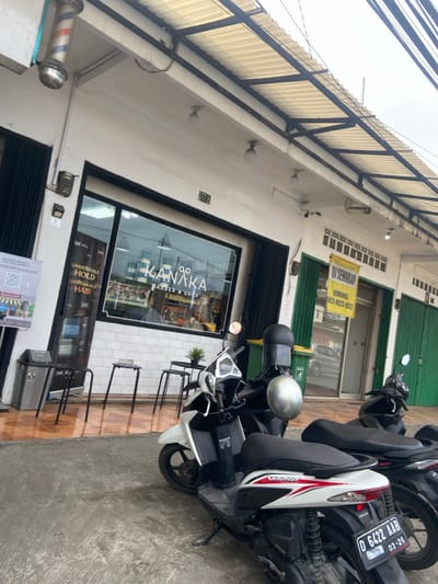 The image shows the exterior of a barbershop called "GO KANAKA BARBER & SHOP" in Bojongsoang, Indonesia. The barbershop has a large storefront window with a sign that reads "GO KANAKA BARBER & SHOP". There are three small tables with chairs in front of the storefront window. Three motorcycles are parked in front of the shop. The nearest motorcycle is white with black and red accents. The middle motorcycle has a black body. The last motorcycle is partially visible in the image and has a black body. The motorcycles are parked in front of the barbershop, which is located on a street. The street is paved with concrete and has a sidewalk. The sidewalk is empty. The barbershop is located in a commercial area. There is a sign in the window that reads "UNBREAKABLE HOLD UNBREAKABLE HAIR". There is a green sign to the right of the barbershop. There is a red sign to the left of the barbershop, which includes QR codes. There is an automatic door just to the left of the sign. There is a large metal pole attached to the roof of the barbershop, which is hanging down to the ground. It appears to be a barber pole. The street is wet and there are puddles of water on the ground. There is a white building next to the barbershop. There is a metal canopy over the street and sidewalk. The image is taken from a street-level perspective. The image is taken on a cloudy day. The location is -6.971, 107.637.
