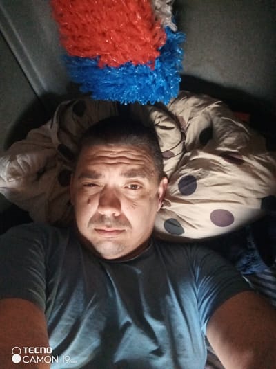 A close-up, high-angle selfie photograph depicts a male individual lying supine in an indoor setting. The man, appearing to be middle-aged, has short dark hair, a light mustache, and visible facial lines, particularly on his forehead. He is looking directly into the camera, with his left eye appearing slightly narrower than his right. He wears a short-sleeved, light blue or grey t-shirt. To the right of his head, a beige fabric with large dark brown/purple polka dots is visible, consistent with a pillow or bedding. Directly above the man's head, in the upper portion of the frame, are two clustered, fluffy decorative objects made of crinkled plastic-like material; the top layer is bright red, and the layer beneath it is bright blue. The surrounding background is dark and indistinct, suggesting an enclosed space. The image displays a "TECNO CAMON 19 Neo" watermark in the bottom left corner. The specific location within the city of Pridorozhnyi, Russia, cannot be determined from the visual evidence.