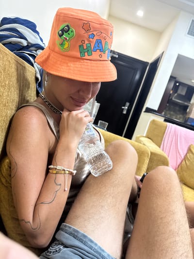 A person is seated on a yellow couch, wearing an orange bucket hat with a dinosaur design and the word "HAPPY". The person is holding a water bottle and has tattoos on their arm. The background shows a room with doors and what appears to be a kitchen area. The image is taken indoors in what seems to be a residential setting. The location is the city of Al Fiqait, United Arab Emirates.