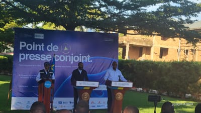 The image shows a joint press conference in an outdoor setting. A blue banner serves as a backdrop, featuring logos of health organizations and the phrase "Point de presse conjoint," which translates to "Joint press conference." Three men stand at podiums, seemingly giving speeches. The backdrop indicates a collaboration between the Government of the Democratic Republic of Congo and the World Health Organization, with a focus on the "Situation de la riposte la Maladie à virus" which translates to "Response to Viral Diseases situation". The event seems to focus on Ebola, as the podiums have "Ministre de la Communication Briefing - Riposte contre Ebola" and "Ministre de la Santé Publique Briefing - Riposte contre Ebola" on them, indicating a media briefing by both the Minister of Communication and Minister of Public Health to discuss the Ebola response. The setting is specified as Ituri, with the date being May 30, 2026. Logos of various international aid organizations are visible at the bottom of the banner, including CDC, RTI, and IFRC. In the foreground, the heads of audience members can be seen. A building under construction, obscured by a large tree and dense greenery, is visible in the background. The watermark "Joseph Kisuki" is in the bottom right corner of the photo. From the sign on the podiums, the event is taking place in Ituri, a province of the Democratic Republic of the Congo, and likely in the city of Bunia.