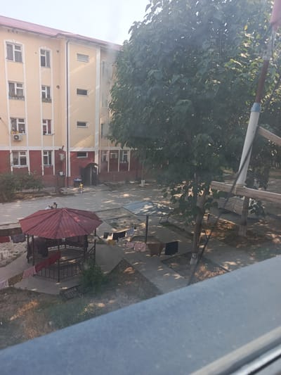 In the city of Hakkulabad, Uzbekistan, an outdoor courtyard is depicted. A multi-story building with beige and red facades, featuring numerous windows and air conditioning units, forms the background. In the foreground, a red-roofed gazebo stands, surrounded by a metal fence. Laundry hangs on clotheslines draped around the gazebo and on the ground nearby. A person is seated inside the gazebo. Elsewhere in the courtyard, a young child in a yellow shirt is visible near the building's entrance, and another person is seated on a bench. Trees with dense foliage are scattered throughout the scene, casting shadows on the paved ground. A tarp-covered object, possibly a vehicle, is situated to the right. The overall impression is of a residential area with everyday activities taking place outdoors.