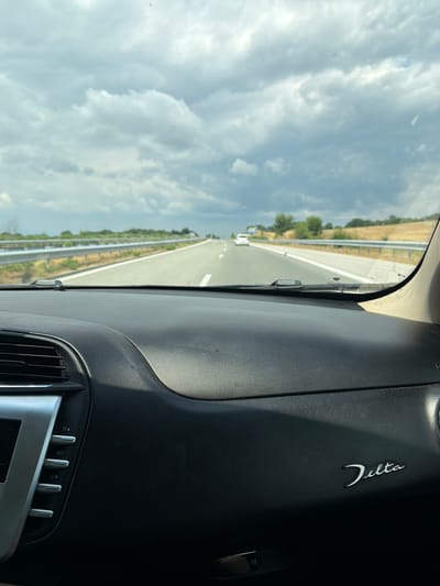 The image shows a view from inside a car dashboard looking out at a highway. The dashboard is black and has a silver trim and a "Delta" emblem. The highway stretches into the distance with a guardrail on the left. The sky is cloudy and the weather appears overcast. There are trees and fields visible on either side of the highway. The scene suggests a road trip or travel on a highway. The location is the city of Aleksandrovo, Bulgaria.