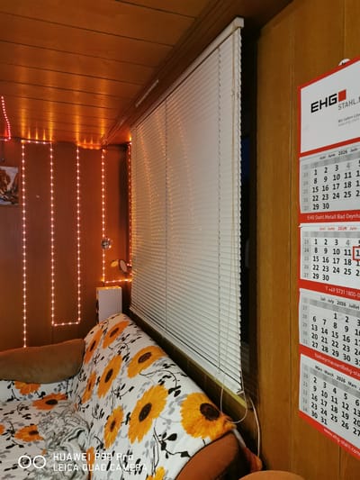 The image shows an indoor scene, likely a room, with a couch covered in a sunflower-patterned throw blanket. The room is decorated with string lights that create a warm, ambient glow. A set of white blinds covers a window. On the right side of the image, there are two calendars, one for June-July 2026 and another for March 2026. The calendars are for EHG Stahl, which has an address in Bad Oeynhausen, Germany. A phone number and website address are also visible on the calendars. The presence of these calendars suggests the location is either a business or a home where EHG Stahl products or services are relevant. The overall impression is one of a relaxed, personalized space with functional elements.