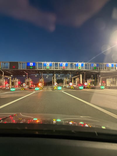 The image shows a toll plaza at dusk on a highway. There are multiple lanes with toll booths. Overhead signs display traffic signals, with some lanes showing a red X and others a green arrow, indicating open or closed lanes. Several cars are visible in the lanes, approaching or at the toll booths. The road is wet, reflecting the lights from the toll plaza. The sky is dark blue with some clouds and a bright light source is visible on the right side of the image, possibly a street lamp. The scene is located on the A4 motorway near Mysłowice, Poland.