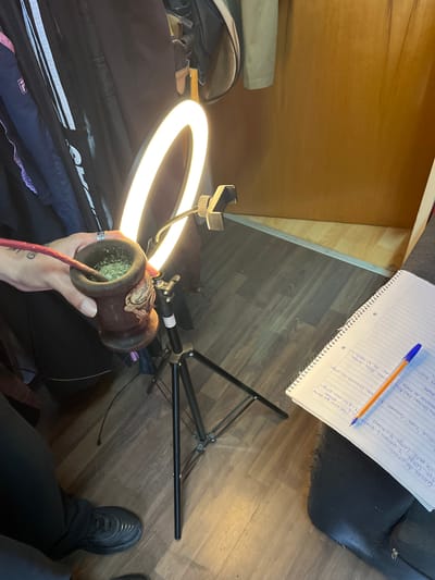 The scene appears to be an indoor setting in Llavallol, Argentina, showing a person preparing what seems to be mate, a traditional South American caffeinated drink. The person's hand holds a gourd-shaped container filled with mate leaves, and a metal straw is inserted. A ring light is positioned to illuminate the subject. A notebook with handwritten text and a pen, possibly containing notes in Spanish including references to "clases de patin" (skating classes), sits on what looks like a cushion. The floor is visible, and part of a shoe is present. A garment bag or bag with white stripes is also in view.