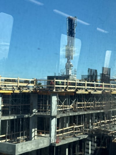 This image depicts an active multi-story building construction site in Hialeah, United States of America, viewed from an elevated perspective through a reflective glass pane. The primary subject is a concrete structural frame in various stages of completion, extending upwards through multiple levels. Extensive wooden formwork and scaffolding support the concrete slabs and columns on the visible floors. On the uppermost completed deck, several sections of vertical rebar cages are visible, indicating areas awaiting concrete pours. A large, articulated concrete pump boom extends vertically from a central point on the structure, its arm retracted. Three construction personnel are present on the top deck; one wears an orange high-visibility vest, and at least two others wear green or yellow high-visibility vests. They appear to be engaged in supervisory or operational tasks on the unfinished floor. The sky overhead is clear and bright blue. Distant palm trees are faintly visible on the horizon, consistent with the geographic location.