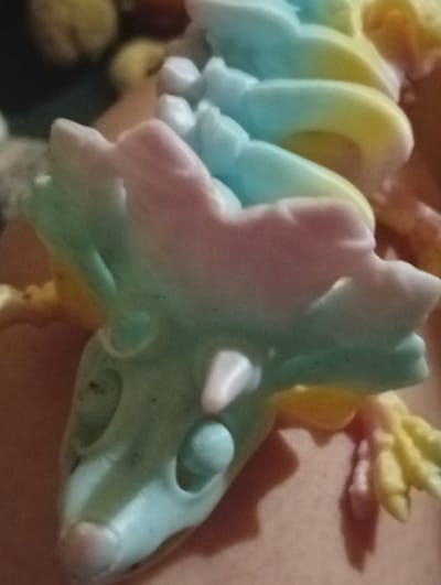 The image shows a close-up, top-down view of a colorful, possibly soap or candy, lizard figurine. The figurine exhibits a gradient of pastel colors, including pink, light blue, and yellow, along its segmented body. Some parts of the figurine appear translucent. The background is blurry and dark, with hints of other colorful objects. There is no discernible information about the city of Berezivka, Ukraine, or any human activity within the image. The focus is entirely on the detailed texture and coloration of the lizard figurine.
