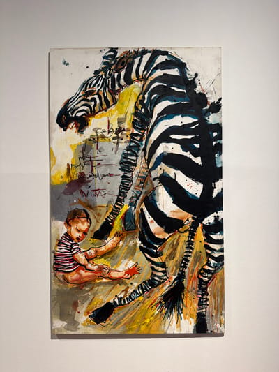 This is an image of a painting on a wall. The painting depicts a large zebra and a baby. The zebra is rendered in black and white stripes with bold brushstrokes, with its head down and mouth open. A baby, wearing a striped shirt, is sitting on the ground to the left of the zebra, looking down. The background of the painting is abstract with splashes of yellow, grey, and red. The painting is hanging on a textured white wall. The context of La Cuesta, Spain, is not discernible from the image itself.