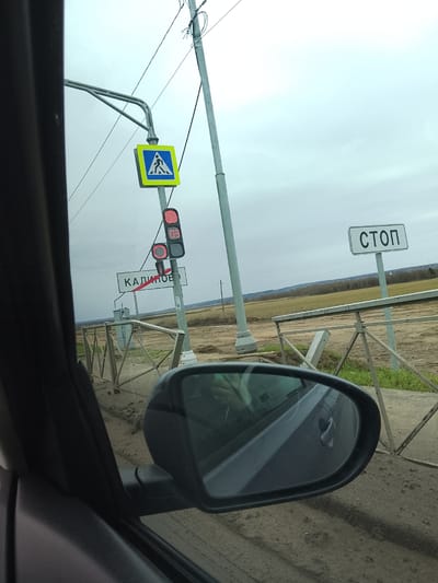 The image shows a view from inside a car in the vicinity of Kalinovo, Russia. Outside the car window, there is a rural landscape with fields and a slightly overcast sky. Roadside infrastructure is visible, including a "STOP" sign and a sign indicating the location as "Kalinovo." There is a pedestrian crossing sign and traffic lights with red lights illuminated. A metal fence lines the edge of the road. The car's side mirror is visible in the foreground.