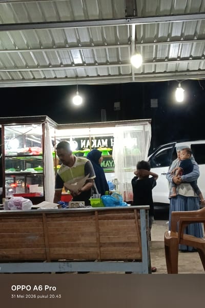 Here is a forensic description of the image:
The image shows a nighttime scene in what appears to be an outdoor food stall or small restaurant, possibly in Sangso, Indonesia. The timestamp indicates May 31, 2026, at 20:43.
In the foreground, a man wearing a striped polo shirt stands behind a wooden counter, appearing to prepare food or handle items. To the left, there's a glass display case containing food items.
To the right, there is a woman in a blue hijab and a woman in a long blue dress holding a child. A younger child stands in front of them with their hands covering their eyes. A white vehicle is visible in the background.
The area is lit by several hanging light bulbs under a corrugated metal roof. The ground appears to be unpaved. There is a stack of plastic chairs to the right. A plastic bin with blue contents is visible on the counter, along with a small green basket.