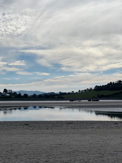 The image depicts a serene landscape with a body of water, possibly an estuary or bay, reflecting the sky. The foreground is a sandy beach with ripples and some debris, indicating it is exposed at low tide. In the mid-ground, the shallow water shows intricate patterns on its sandy bed. Two tractors, one pulling a trailer, are visible on the exposed seabed, suggesting some form of activity. Across the water, a tree-lined shore leads to rolling hills dotted with wind turbines in the distance. A few houses are nestled among the trees on the left side of the shore, and the sky is filled with a dramatic display of clouds. The scene is quiet and expansive, with a sense of natural beauty and human presence. The location is identified as Tullycullion, Ireland.