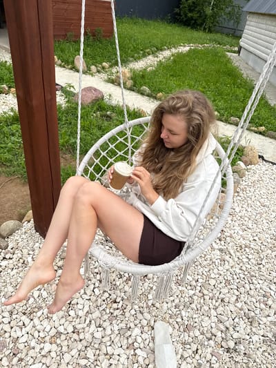 A young woman with long, wavy blonde hair is sitting in a white, woven hanging chair outdoors. She is wearing a white hooded sweatshirt and dark brown shorts. She is holding a small, disposable cup with a lid in her hands and looking down at it, appearing to be taking something out or adding something to it. Her feet are bare and resting on the ground, which is covered in light-colored gravel.

To the left of the chair, there is a grassy area with some rocks and a wooden fence. Behind the grass, there is a building with a grey, corrugated roof and siding. The scene appears to be a backyard or a garden area in Ryzhkovo, Russia.

The lighting suggests it is daytime, possibly overcast or diffused sunlight, as there are no strong shadows. The overall mood seems relaxed and casual, with the woman enjoying a drink in a comfortable outdoor setting. There is no visible text in the image.