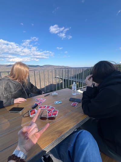 A medium-shot photograph shows a game of Uno being played on a wooden table. The location is outdoors with a view of a mountainous landscape, likely near Tuhovishta, Bulgaria. Two people are visible at the table. One wears a black hoodie and sits opposite a person wearing a dark jacket. Uno cards are scattered on the table, along with a water bottle, sunglasses, and mobile phones. A third person's hand, wearing a watch and rings, is in the foreground making a hand gesture. A metal railing is visible in the background, suggesting a balcony or elevated viewpoint.