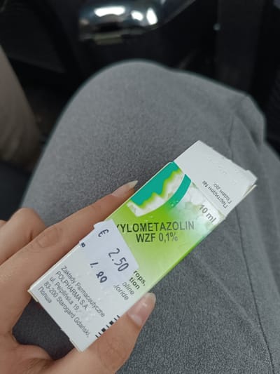 An individual's left hand holds an upright, rectangular white and green box of Xylometazolin WZF 0.1% nasal drops, 10 ml volume. The box displays manufacturer information for "Zaklady Farmaceutyczne POLPHARMA S.A." in Starogard Gdanski, Poland. A price sticker affixed to the box indicates "€ 2.50" with an obscured "1.90" below it. Cyrillic text on the box edge reads "Партиден №" (Batch No.) and "Годен до: 010525" (Expiration date: May 2025). The hand exhibits long, natural-colored, pointed fingernails. The box is positioned over the individual's lap, which is covered in grey fabric, likely trousers. The background is dark and blurred, suggesting an interior space, possibly a vehicle, with indistinct reflective elements. The scene is located in Muselievo, Bulgaria.