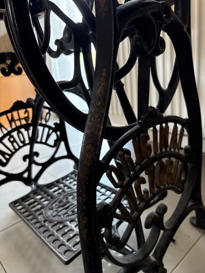 This media file shows a close-up, angled shot of the ornate cast-iron base of an antique sewing machine. The subjects are the intricate metalwork of the machine's stand, which features decorative scrollwork and embossed lettering. The scene is likely indoors, with a soft, diffused light suggesting a bright day filtering through a window or other light source, which is visible as a blurred background. The setting appears to be a home or workshop. There is no discernible activity taking place; the focus is on the static, decorative details of the sewing machine. Notable details include the metallic sheen of the dark cast iron, and faint gold detailing on a horizontal bar, possibly an ornamental pattern. The embossed text on a circular medallion reads "WHITE". The overall impression is one of historical craftsmanship and vintage aesthetics.