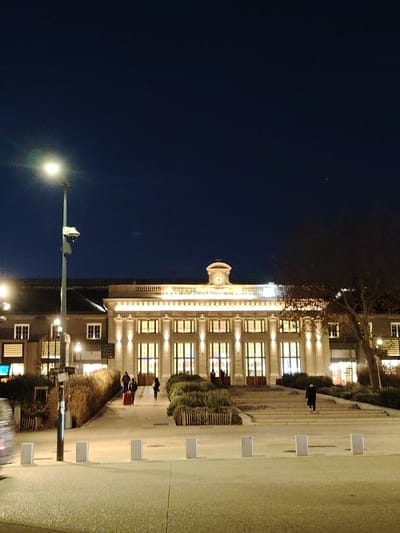 An illuminated building at night features a central clock tower and several arched windows. People walk up steps to enter the building. Bushes and low barriers flank the steps. A streetlight stands on the left, and a tree is visible on the right. The foreground includes a paved area with short, white bollards. The sky is dark, and the scene is lit by artificial lights.