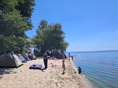 On a sandy beach in Maksymivka, Ukraine, a group of people are enjoying a sunny day by a large body of water. Tents are set up on the sand, suggesting a camping or overnight stay. Several individuals, including children, are visible on the beach and in the shallow water. Some are playing in the sand, while others are wading or standing near the shoreline. The water appears calm and blue, reflecting the clear sky. Trees line the back of the beach, providing shade. The overall scene depicts a recreational gathering in a natural outdoor setting.