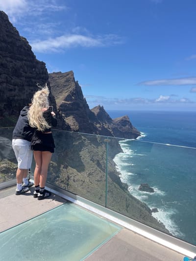 The image depicts a couple standing on a glass platform overlooking a coastal landscape. The woman has long, blond hair, wears a black long-sleeved shirt and black skirt and black Adidas sneakers with three white stripes, and is holding a cell phone with both hands, potentially taking a photo. The man is wearing a blue denim jacket and white shorts with white socks and black Adidas sneakers with three white stripes. They appear to be standing very close to one another. The observation deck is on the Acantilado del Balcón in La Aldea de San Nicolás, Spain. The view shows dramatic, rocky mountains that plunge into the blue ocean, and white waves crash at the base of the mountains. The sky is bright blue with some scattered clouds. A glass barrier surrounds the platform. The ground appears to be concrete.