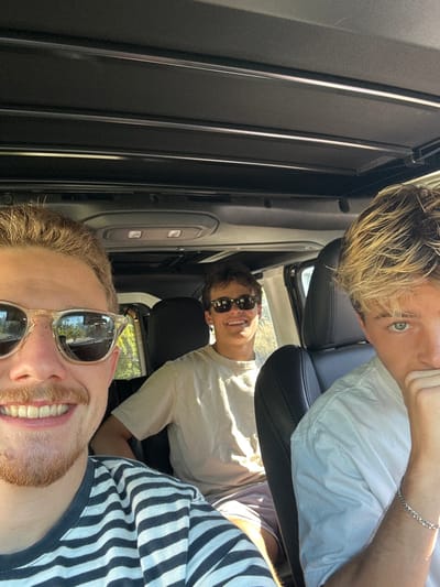 This image depicts three young adult males inside a vehicle, located in Summerland, United States, under natural daylight.
In the foreground left, a male with light brown hair, a mustache, and a short beard wears clear-framed sunglasses. He is smiling, showing teeth, and dressed in a horizontal striped t-shirt. Reflections visible in his sunglasses show indistinct architectural or structural elements.
Behind the first male and centrally positioned, a second male with short, dark hair is visible. He wears dark sunglasses, a light-colored t-shirt, and light-colored shorts. He is smiling and looking towards the camera.
On the right side of the frame, a third male with blonde hair is partially depicted. He wears a plain white t-shirt and a silver link bracelet on his left wrist. His left hand is raised, covering the lower portion of his face, as he looks towards the camera.
The vehicle's interior features a dark, segmented overhead panel structure. All three individuals appear to be occupants of the vehicle.