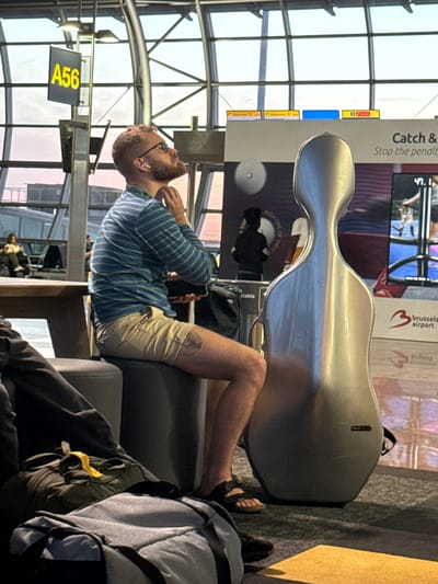 A man with a beard, glasses, and earbuds is sitting in an airport terminal in Zaventem, Belgium. He is wearing a blue and green striped t-shirt and khaki shorts, and has a tattoo on his left thigh. He is looking upwards and touching his neck. Next to him is a large, silver cello case. In the background, there are airport signs, departure boards, and a window showing the sky. Several pieces of luggage are visible on the floor.