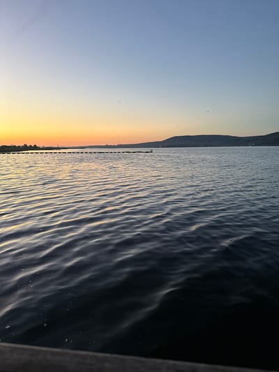 This image depicts a serene body of water with gentle ripples under a twilight sky. The sky transitions from a soft orange glow near the horizon, likely from a sunset, to a pale blue overhead. In the distance, a low, dark landmass, possibly hills or mountains, forms the silhouette against the sky. A long, thin structure, appearing to be a pier or a line of buoys, stretches across the water. A small, dark object, possibly a boat or a distant structure, is visible on the water near the pier. A few small, dark specks in the sky suggest distant birds. The foreground is dominated by the dark, reflective surface of the water, with a wooden edge visible at the very bottom of the frame. There are no people or discernible activity captured in the scene. The overall impression is one of tranquility and the peacefulness of dusk. The location, Казашко, Bulgaria, suggests this is a scene of the Black Sea coast or a nearby large body of water at sunset.
