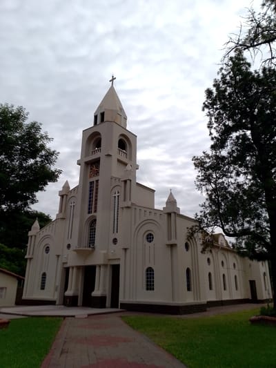 The image depicts the Immaculate Conception Church in Villa Hayes, Paraguay. The church is a light-colored building with a prominent central tower topped with a cross. The tower features arched openings and a clock face. The architectural style incorporates rounded arch windows and decorative vertical elements. The church is flanked by trees on both sides. A paved pathway leads to the church entrance, with grass lawns on either side. The sky is overcast with a layer of clouds. A small building is visible to the left of the church.