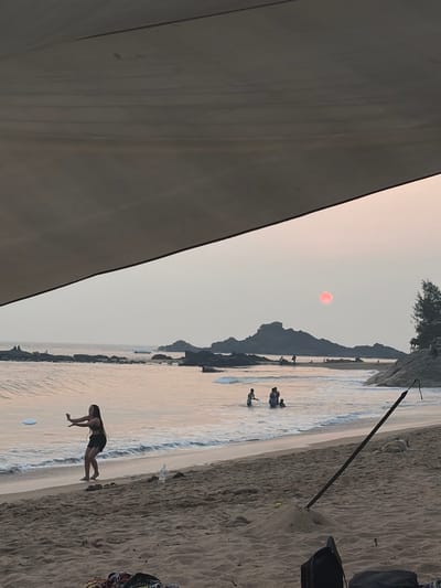 The image captures a beach scene in Gokarna, India, during what appears to be either sunrise or sunset, viewed from beneath a light-colored canopy that diagonally covers the top portion of the frame. The sky is hazy, displaying a prominent, circular, red sun positioned centrally above the horizon. The ocean features gentle waves breaking on a sandy shore, and numerous dark, rocky outcrops are visible both near the coast and further out in the water. On the far right, a small group of evergreen trees grows on a rocky embankment.

In the midground, a female individual, dressed in a dark two-piece swimsuit, stands in the shallow water at the water's edge on the left. She is actively engaged in play, with her arms raised and a white frisbee airborne near her left hand. To her right, further out in the water, a cluster of at least three individuals is visible, partially submerged and wading.

The immediate foreground, within the shaded area under the canopy, consists of sand. Visible objects include dark bags or clothing items, a black strap or pole extending diagonally from a small mound of sand, a yellow tennis ball resting on this mound, and a white plastic bottle lying near the water's edge. A light-colored dog is observed sleeping on the sand in the far right foreground, near the rocky bank.