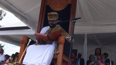 A man, wearing a leopard print hat and a white robe with a leopard print stole, sits on a throne in a large tent in Rukoko, Democratic Republic of Congo. The throne has a wooden lion's head on the armrests. The man speaks into a microphone, and holds a large, closed, red book in his lap. He sits in the foreground, and many people are seated behind him in rows. The people are dressed in a variety of clothing, including suits, dresses, and traditional African attire. The tent is made of white canvas and there are white curtains lining the sides. The scene is likely part of a ceremony or public address.