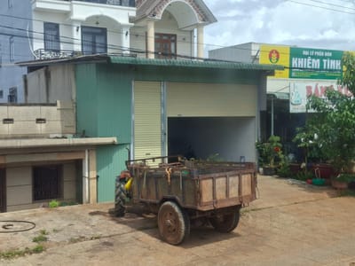 In Đại Ninh, Vietnam, an abandoned trailer is parked in front of a light green corrugated metal building with a partially open rolling shutter door. The trailer is made of weathered wood and rusted metal, with a large, worn tire on the left rear and a smaller, more worn tire on the right rear. A single large tractor tire is visible on the left side of the trailer, suggesting it is a towed unit. The ground is paved with concrete slabs, with patches of sparse vegetation. Behind the trailer, the building appears to be a commercial space, with signage visible further down the street, including a yellow and green sign that reads "ĐẠI LÝ PHÂN BÓN KHIÊM TÌNH." In the background, a modern white building with arched windows and a tiled roof stands prominently. The sky is overcast.