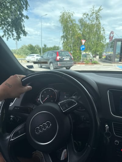 The image is taken from inside a car, looking out through the windshield. A hand with a tattoo reading "09:02" rests on the black leather steering wheel, which prominently features the Audi four-ring logo in the center. The dashboard and instrument cluster are visible, showing a speedometer, tachometer, and various control buttons, some of which read "NAV," "MEDIA," "RADIO," and "INFO." Wipers settings are also visible on a stalk.
Outside the car, the scene depicts an overcast daytime sky. Trees with green foliage are prominent, and a red and white triangular warning sign and a blue circular mandatory sign (indicating a right turn) are visible. Several parked cars, including a dark-colored one and a white one, can be seen, suggesting a parking lot or a street next to a building. Part of a building, possibly a commercial establishment, is also visible. The setting is Radinovo, Bulgaria.